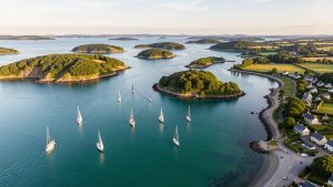 Vue aérienne lumineuse du golfe du Morbihan avec des voiliers, des îlots verdoyants et un village breton au bord de l’eau.