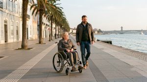 Une femme en fauteuil roulant suit une promenade plane et accessible au bord du Tage à Lisbonne, accompagnée d’un homme, avec des rampes douces et des façades carrelées.
