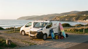 Van aménagé stationné dans un camping autorisé près de Biarritz après une session de surf, avec deux planches posées à côté et l’océan Atlantique en arrière-plan.